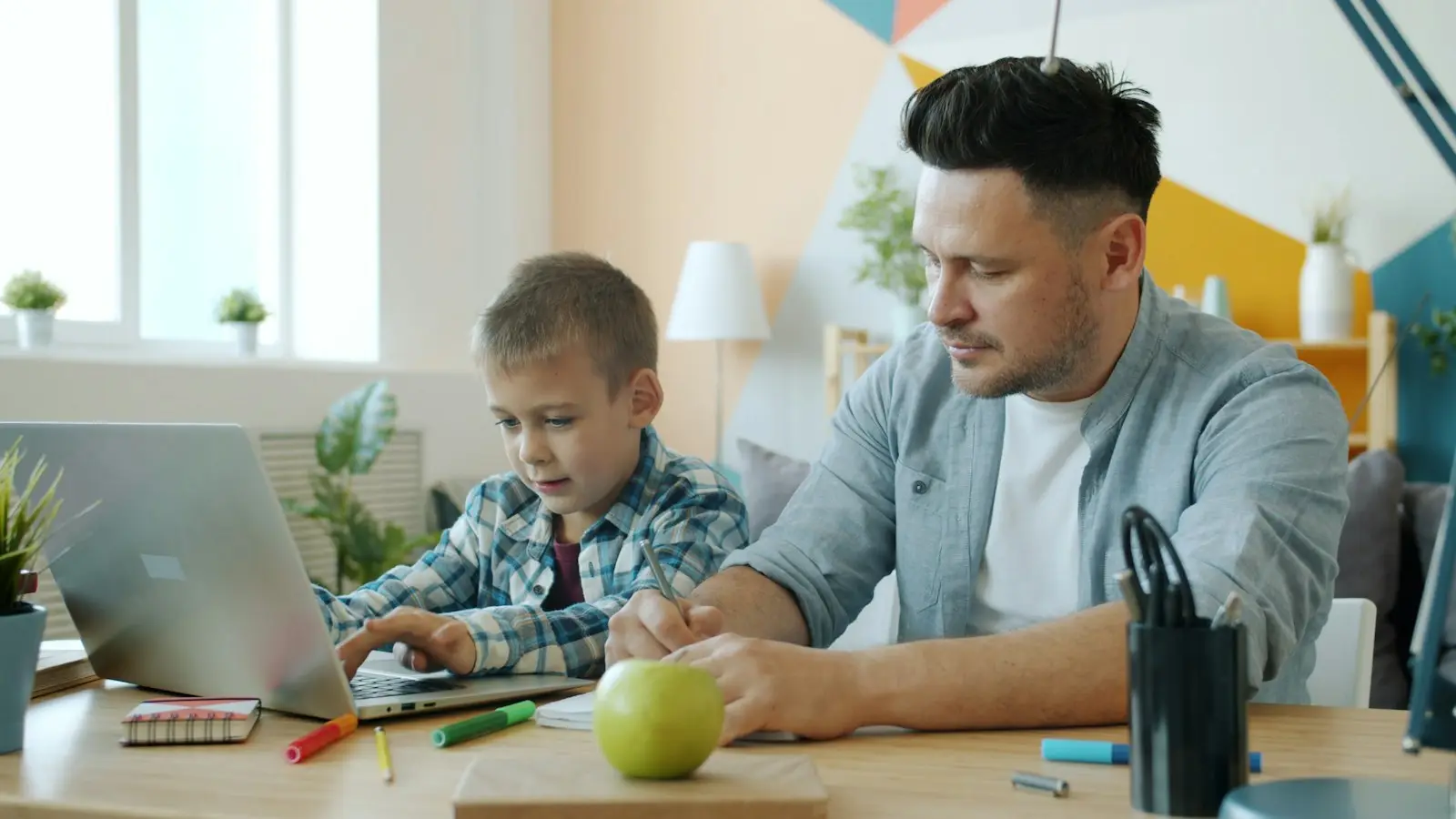 A father and son look at a laptop together at a desk at home.