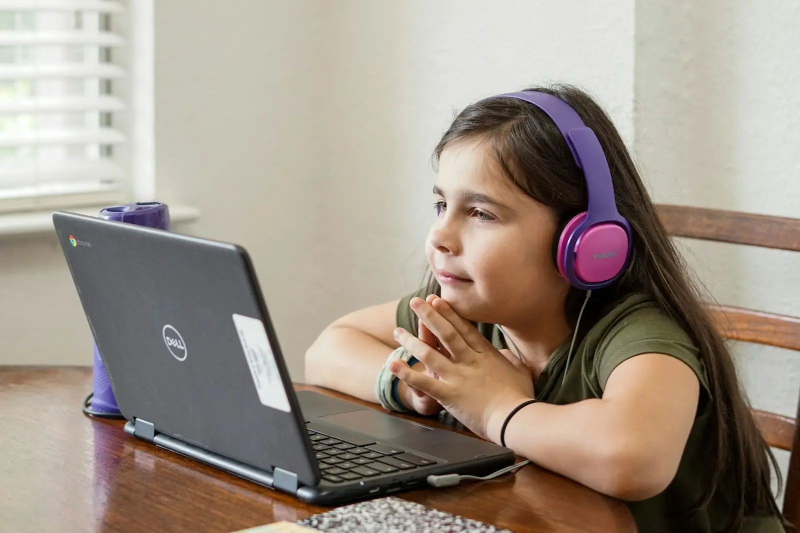 A girl wearing headphones smiles at a laptop during an online lesson at a kitchen table.