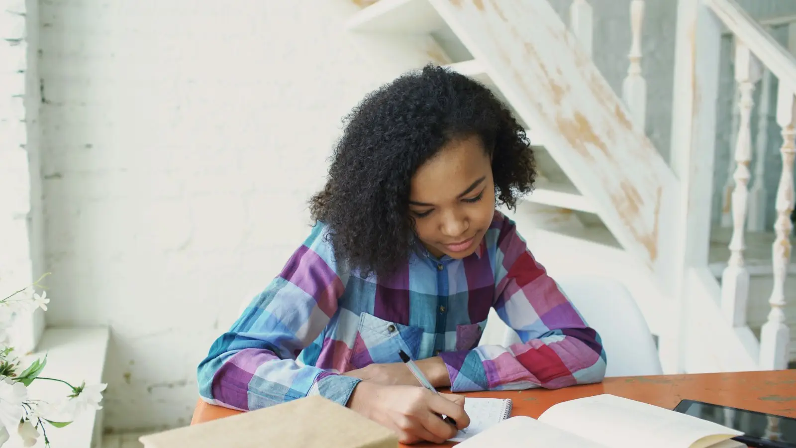 A girl writes in an exercise book at a table by a window.