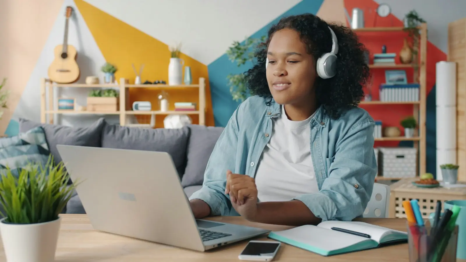 A teenager with headphones works through a problem on a laptop, notebook open beside her.