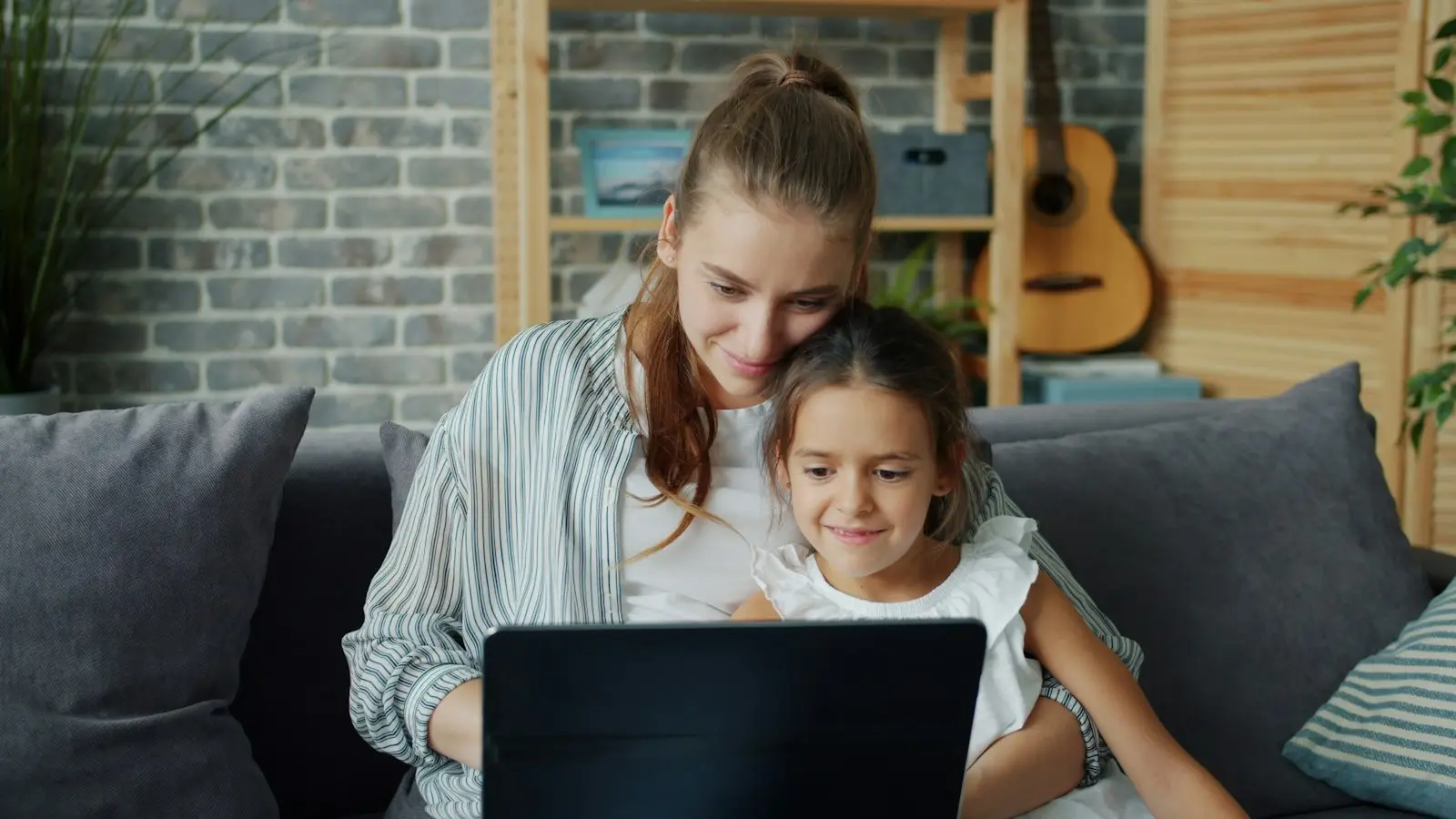 A parent and her daughter read something together on a laptop on the sofa.