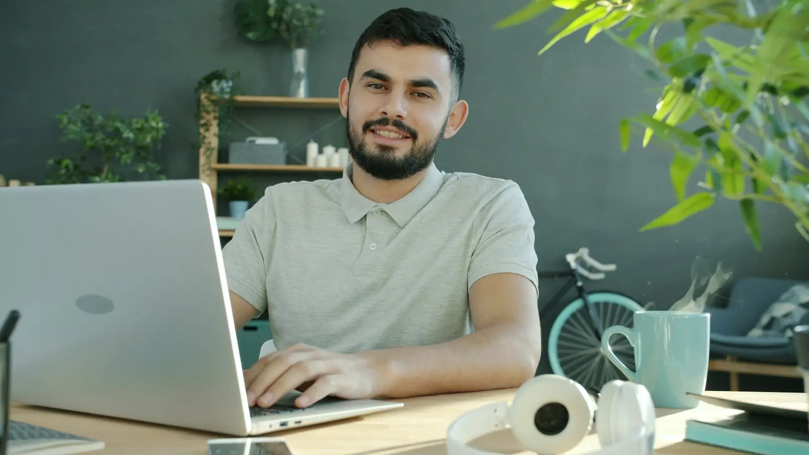 A tutor smiles at his laptop while teaching from a home desk.